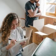 Pensive couple taking notes in bedroom near window while packing stuff in carton box to move into new apartment